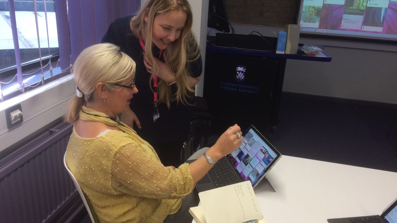 Two ladies (Debbie Baff and Rhian Ellis) in a classroom, Debbie has long blonde hair and a dark top and is leaning towards Rhian who also has blonde hair and glasses, is wearing a yellow top and sitting at a desk pointing at an ipad