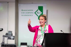 Debbie Baff smiling on stage at the ALT Conference and giving a thumbs up. Debbie is wearing a bright pink cardigan. In the background is a ALT banner sign.